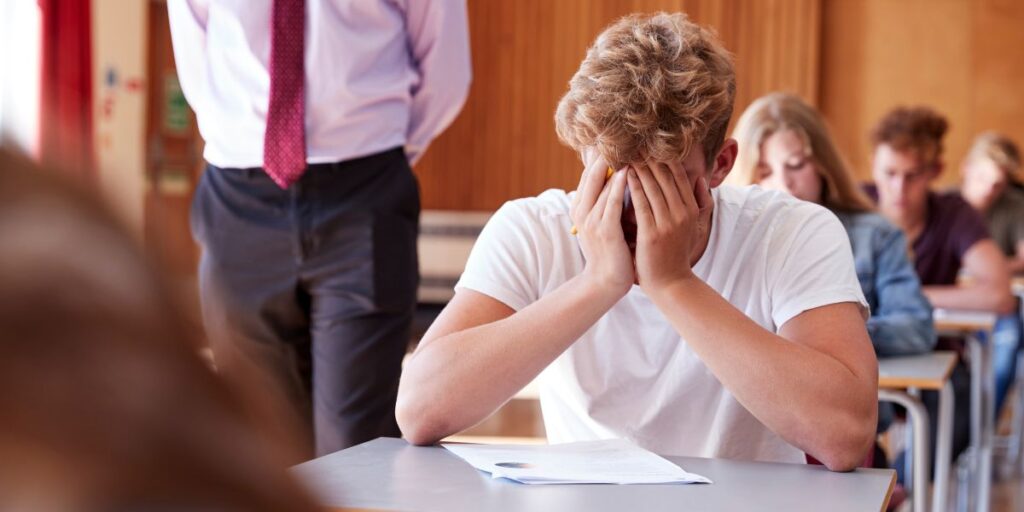 試験会場イメージ。大勢の学生が試験を受ける教室。一人だけ頭を抱えて考え込む学生。すぐ後ろを試験監督が歩くイメージ画像。